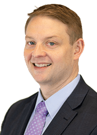 A man with short blond hair wearing a charcoal suit and purple tie smiles at the camera.